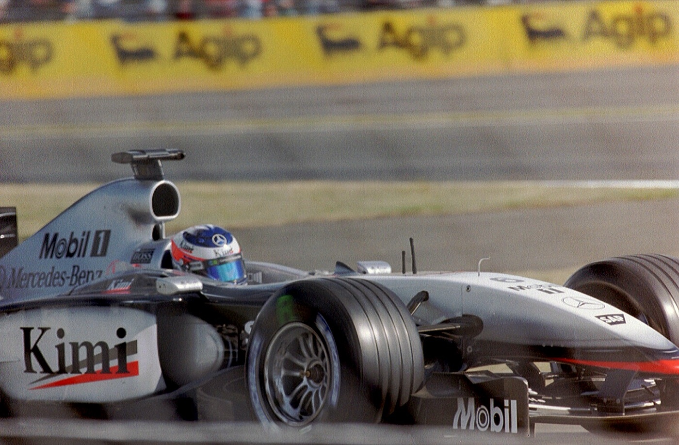 Silver McLaren with Kimi written on the sidepod and a blue and white helmet, passing yellow trackside banners