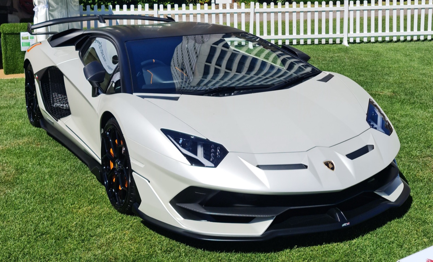 Matt white Lamborghini Aventador SVJ with a black roof and large rear wing, parked on grass in front of a white picket fence