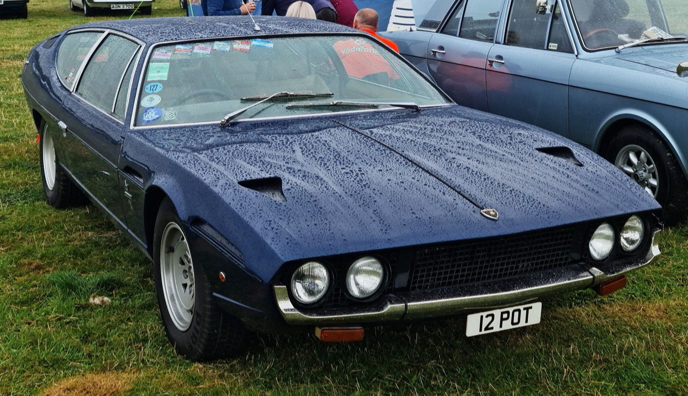 Dark blue 1977 Lamborghini Espada Series III four-seater with four round headlights, beaded with rain on a grass car park