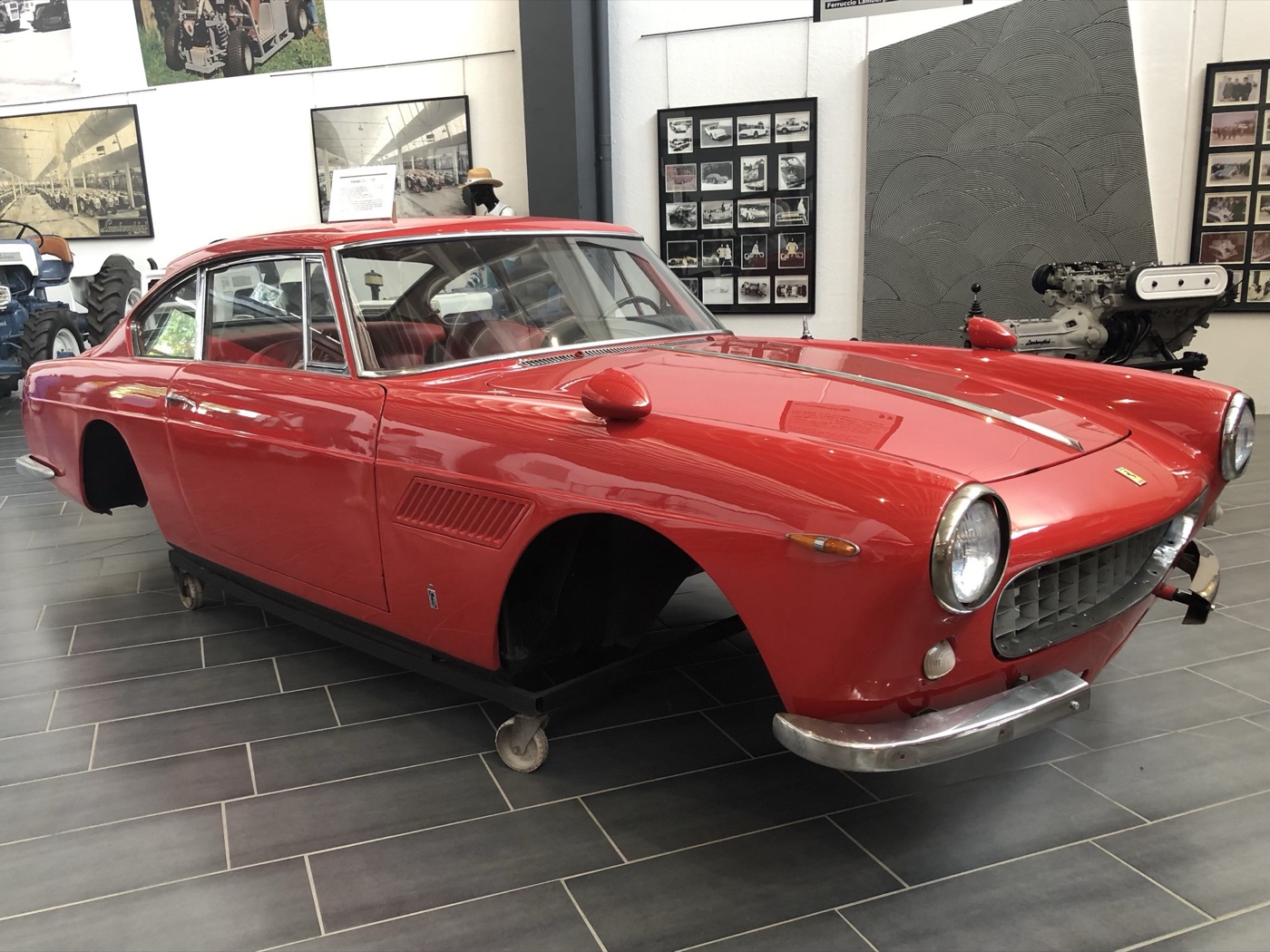 Red Ferrari 250 GTE 2+2 body without wheels on a wheeled dolly in a museum hall, with Lamborghini tractor photographs on the wall behind
