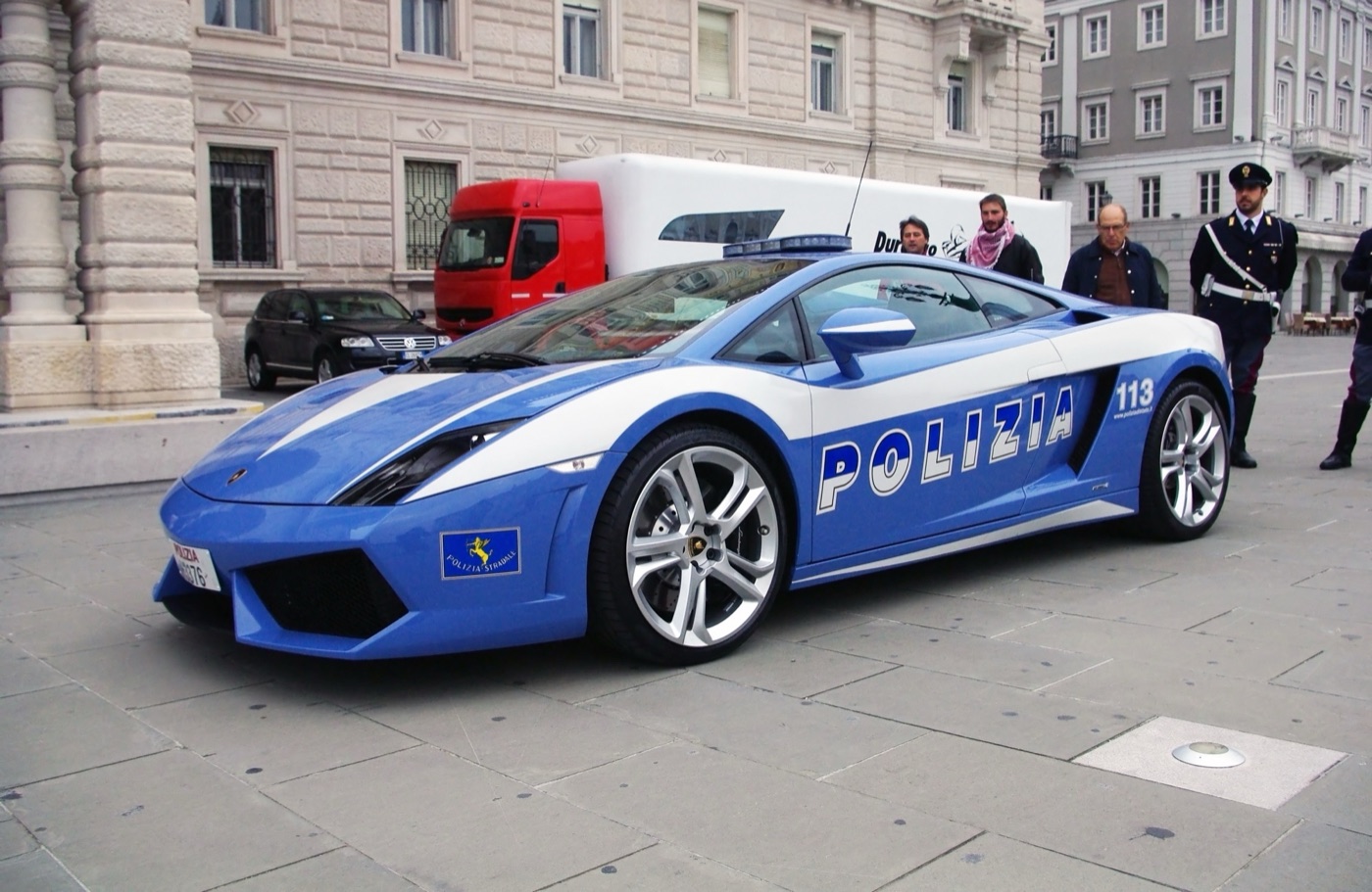 Blue and white Polizia Gallardo LP560-4 parked on a paved square with a uniformed officer standing behind it