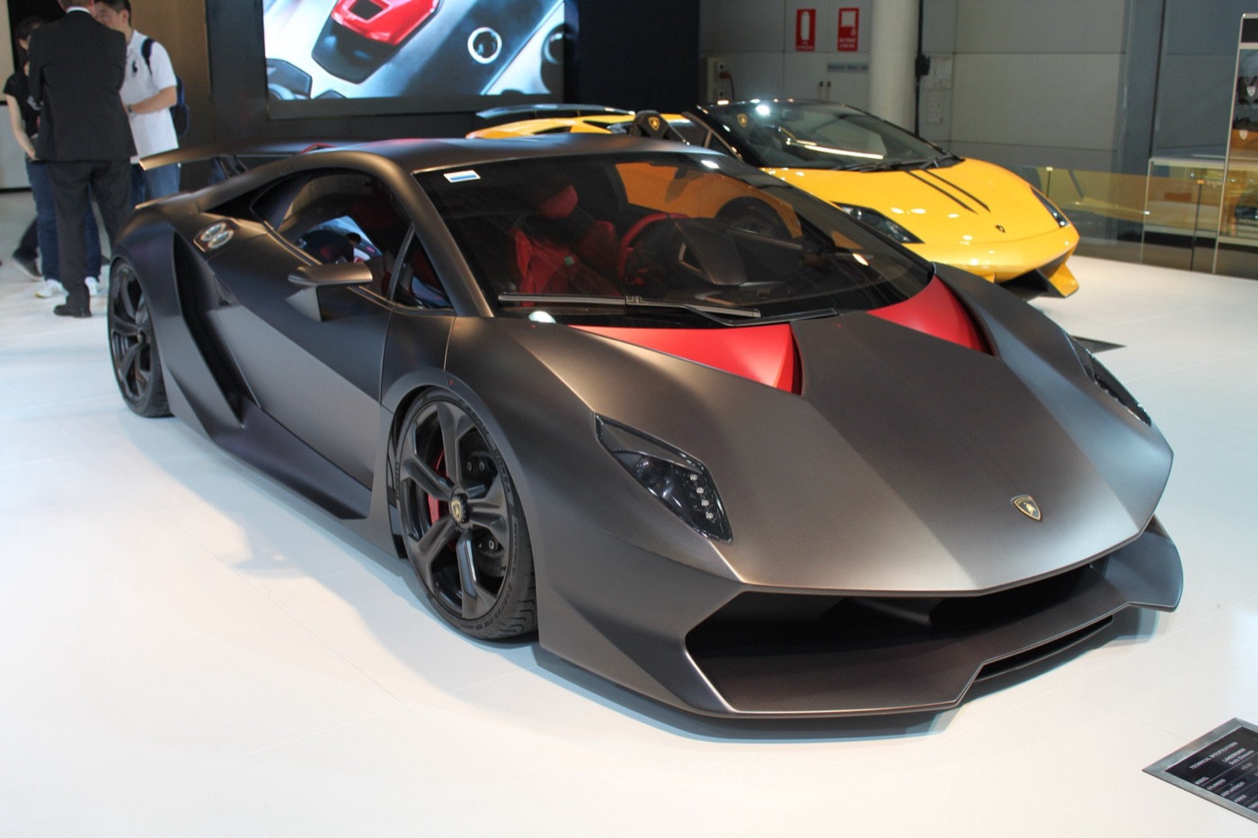 Matt carbon-grey Lamborghini Sesto Elemento with red bonnet vents on a showroom floor, a yellow Lamborghini behind it