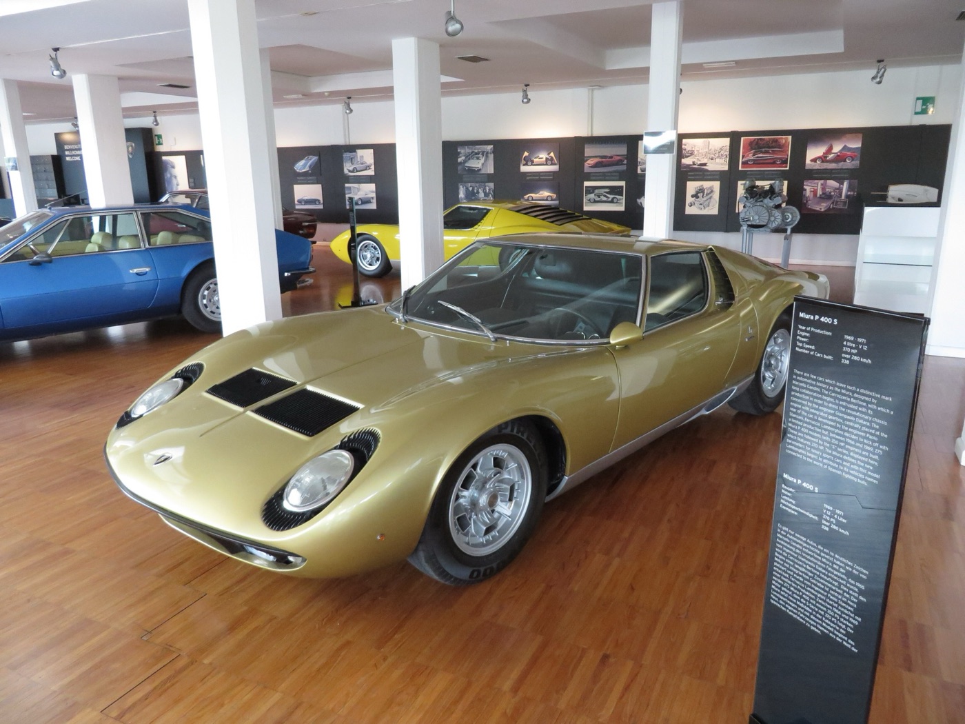 Gold Lamborghini Miura P400 S on a wooden floor inside the Lamborghini museum, with a blue Espada and a yellow Countach behind