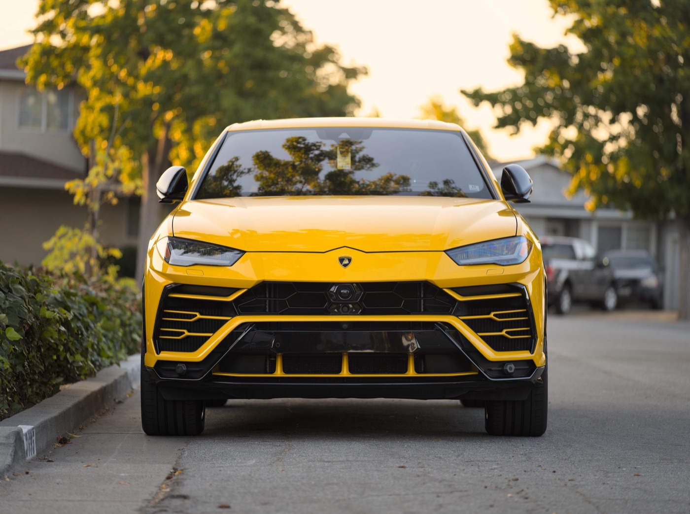 Yellow Lamborghini Urus SUV seen head-on on a suburban street at dusk, with black hexagonal grille inserts