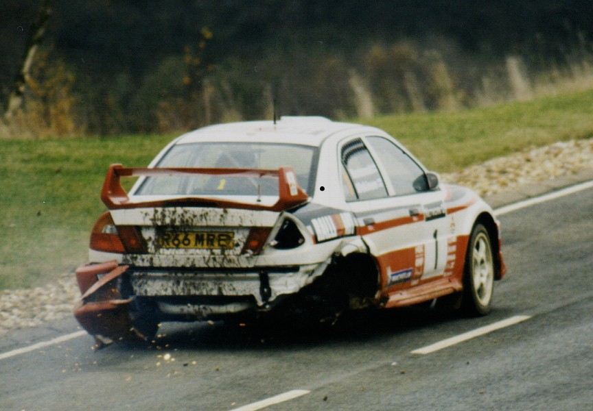 Tommi Makinen’s Mitsubishi Lancer Evo rally car on a tarmac stage with its right rear wheel missing and rear bumper hanging off