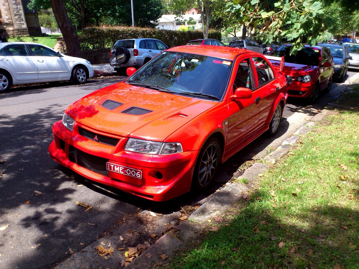 Red Mitsubishi Lancer Evolution VI Tommi Makinen Edition parked at a kerb under trees, with TME 006 number plates
