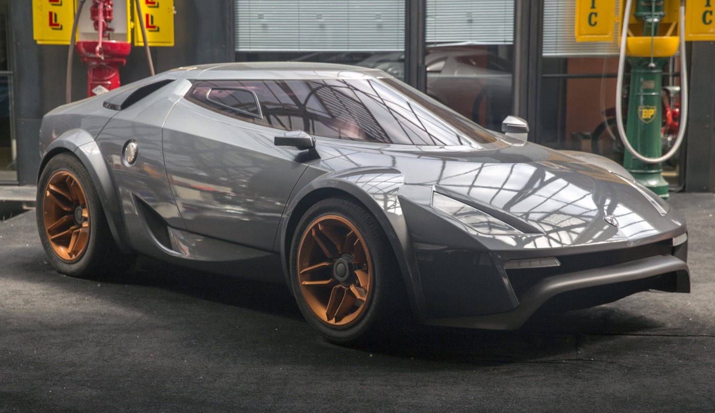 Grey Fenomenon Stratos concept car with bronze wheels and a wraparound windscreen, in front of old petrol pumps