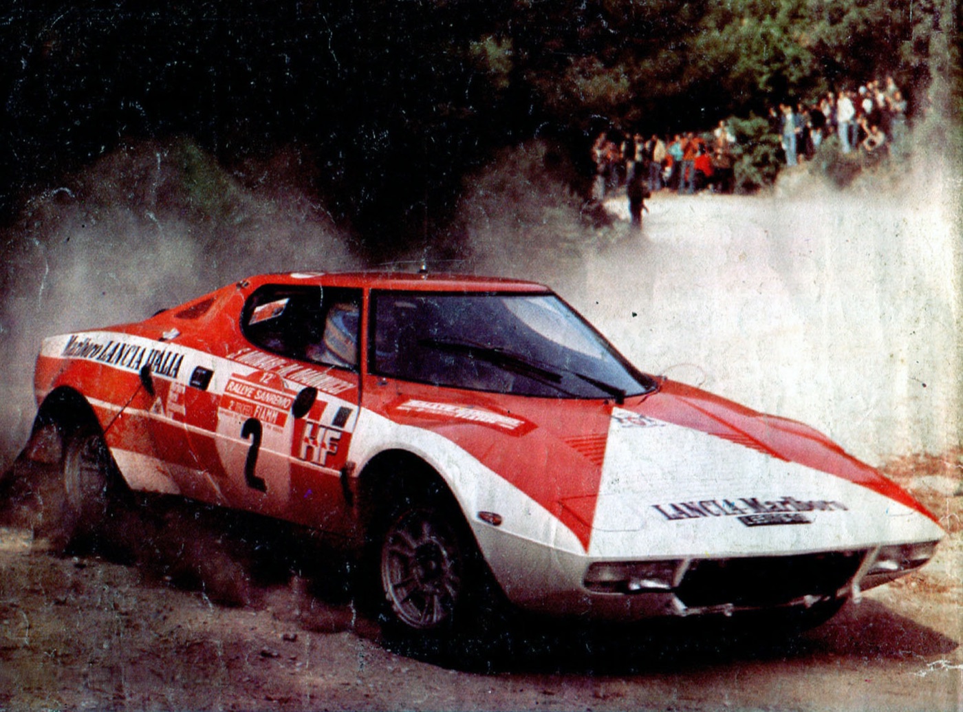 Red and white Marlboro-liveried Lancia Stratos number 2 sliding on a gravel road, with spectators on the bank behind