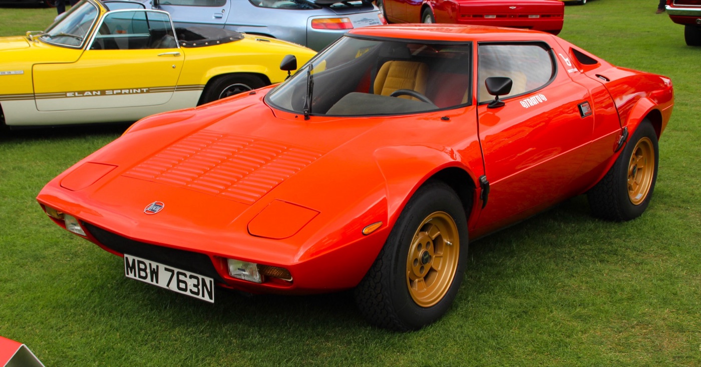 Red-orange Lancia Stratos HF Stradale with gold wheels and a British number plate, parked on grass among other classic cars