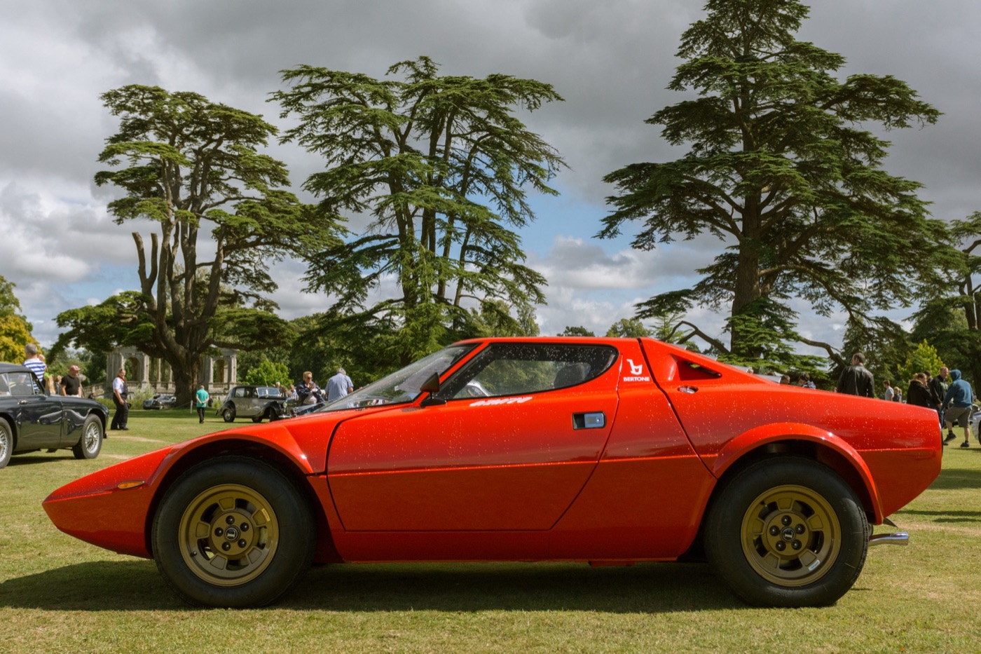 Side view of a red-orange Lancia Stratos Stradale with gold wheels on a lawn, cedar trees behind