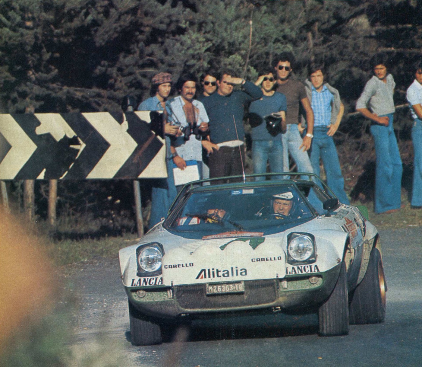 Alitalia-liveried Lancia Stratos rally car on a tarmac bend, with spectators beside a chevron sign