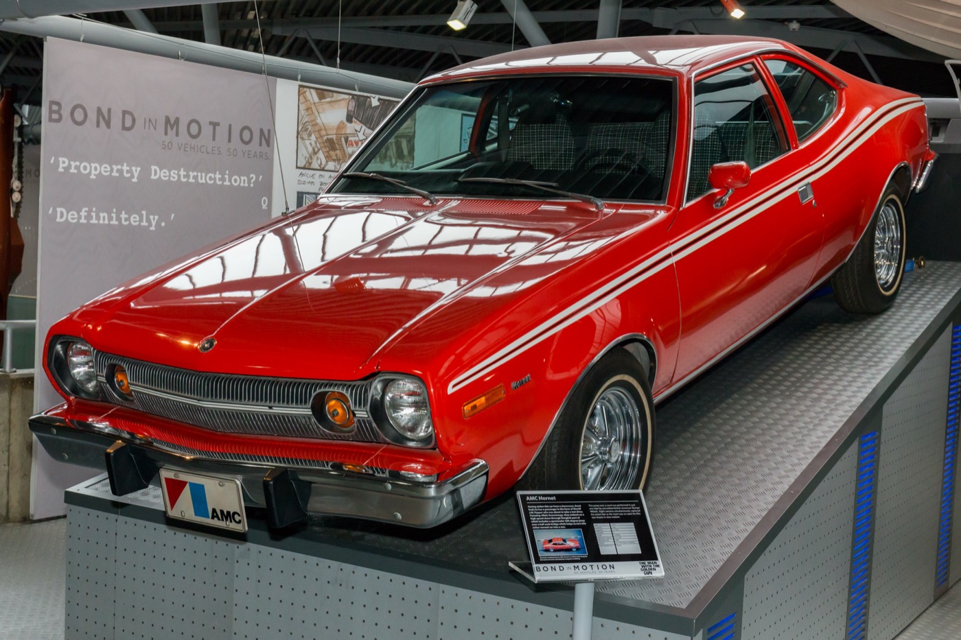 Red AMC Hornet hatchback with white side stripes on a raised stand at the Bond in Motion exhibition