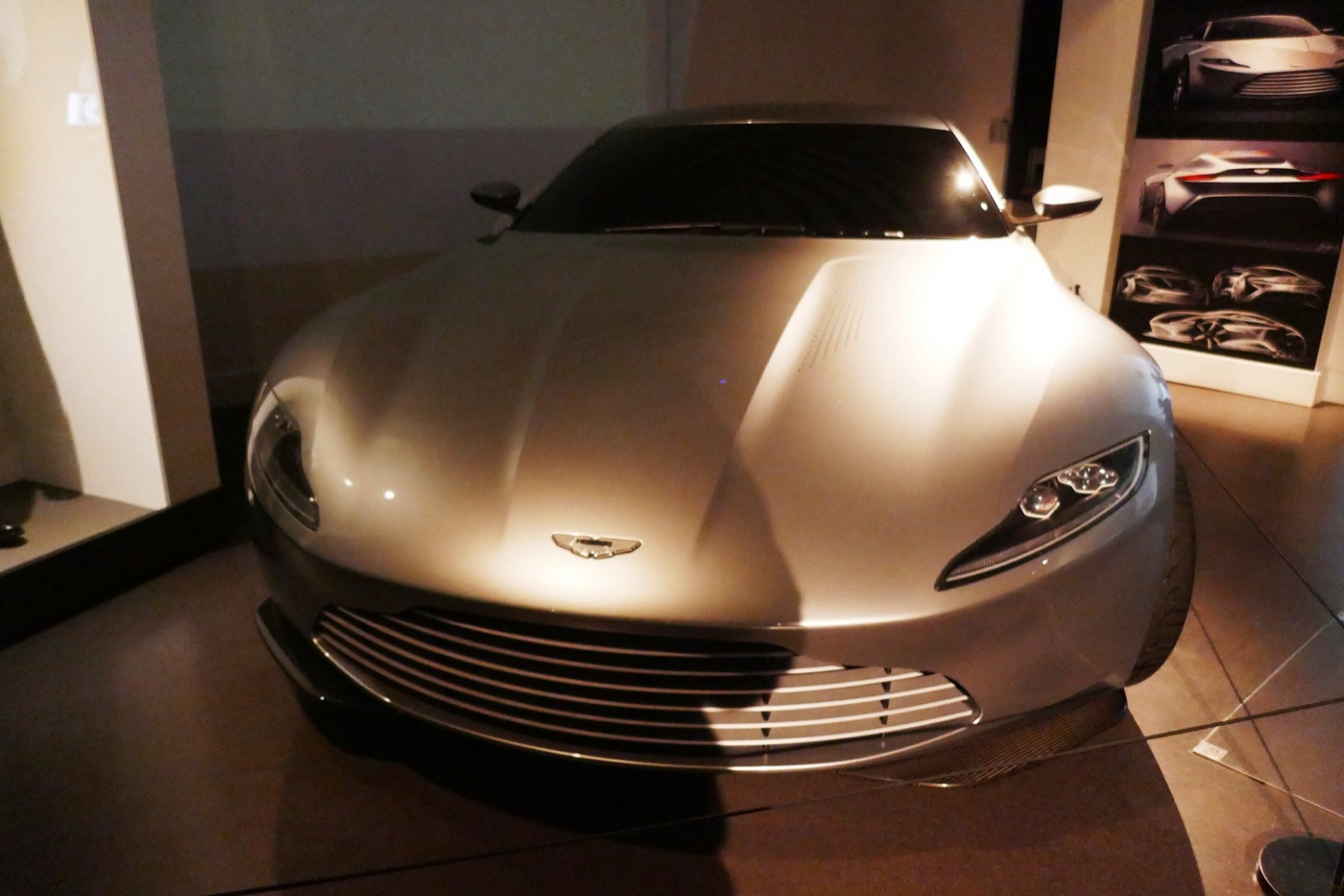 Front of a silver Aston Martin DB10 with its low grille and narrow headlamps, lit in a dark exhibition room