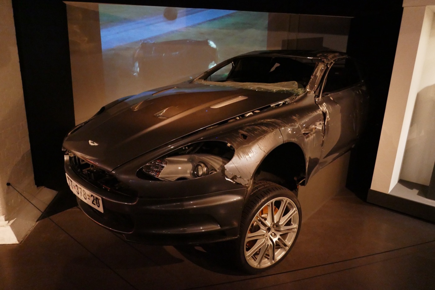 Grey Aston Martin DBS with a crushed roof, missing front wing and torn bodywork, in front of a screen showing the crash