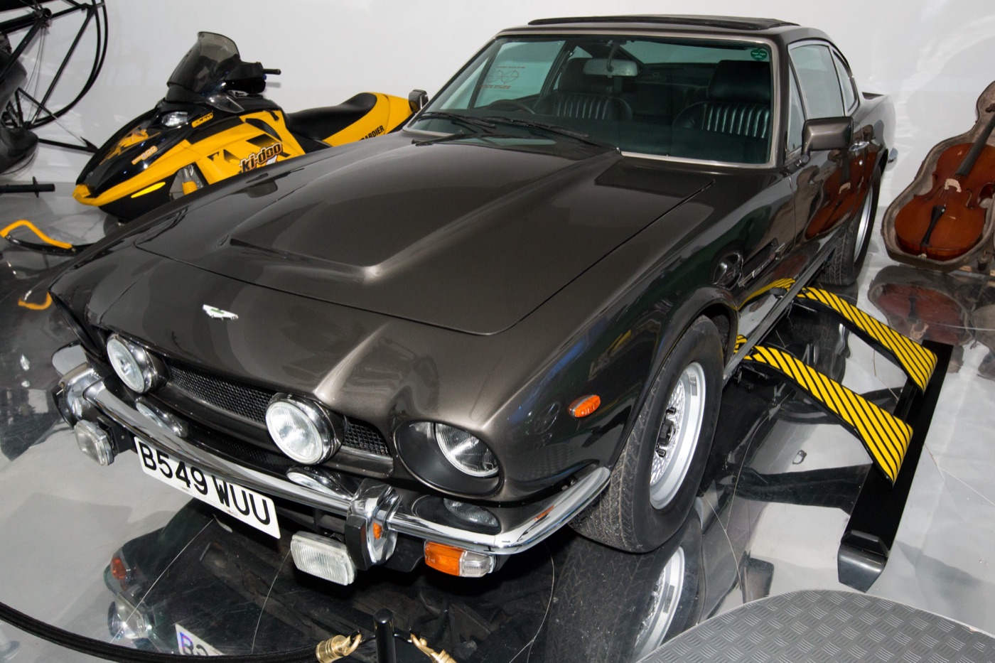 Dark grey Aston Martin V8 with the number plate B549 WUU and its outrigger skis extended, beside a snowmobile and a cello case