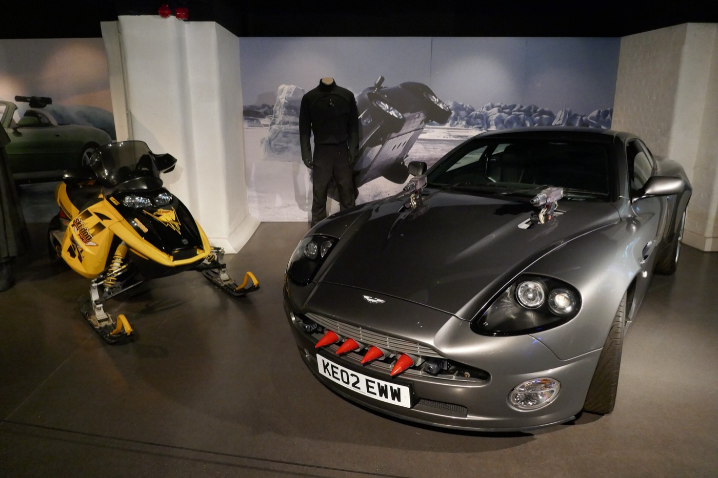 Silver Aston Martin V12 Vanquish with red-tipped missiles in its grille and guns on the bonnet, next to a yellow snowmobile