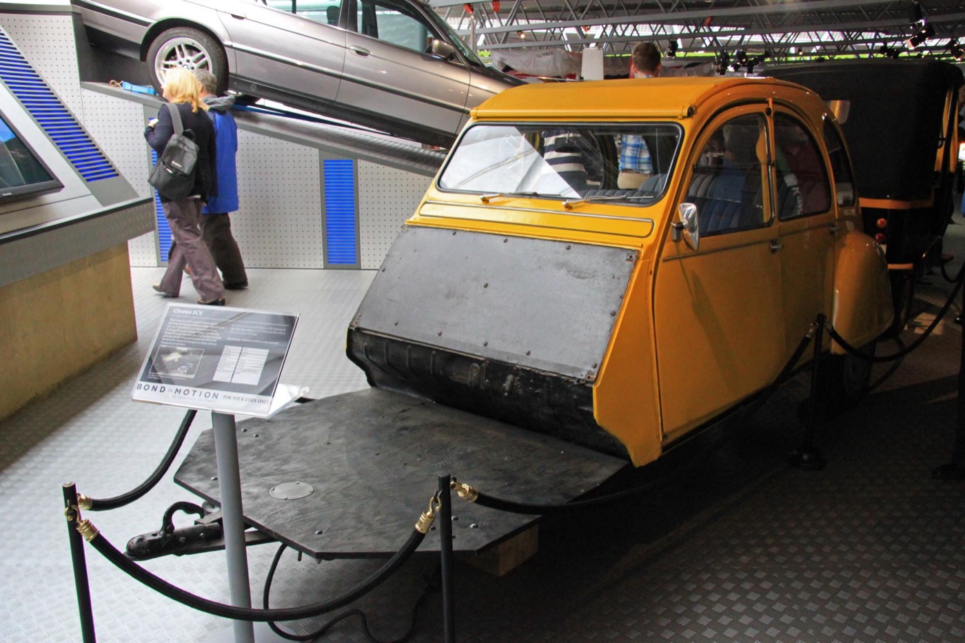 Yellow Citroën 2CV stunt car cut down to its cabin and mounted on a flat steel rig, behind a rope at an exhibition