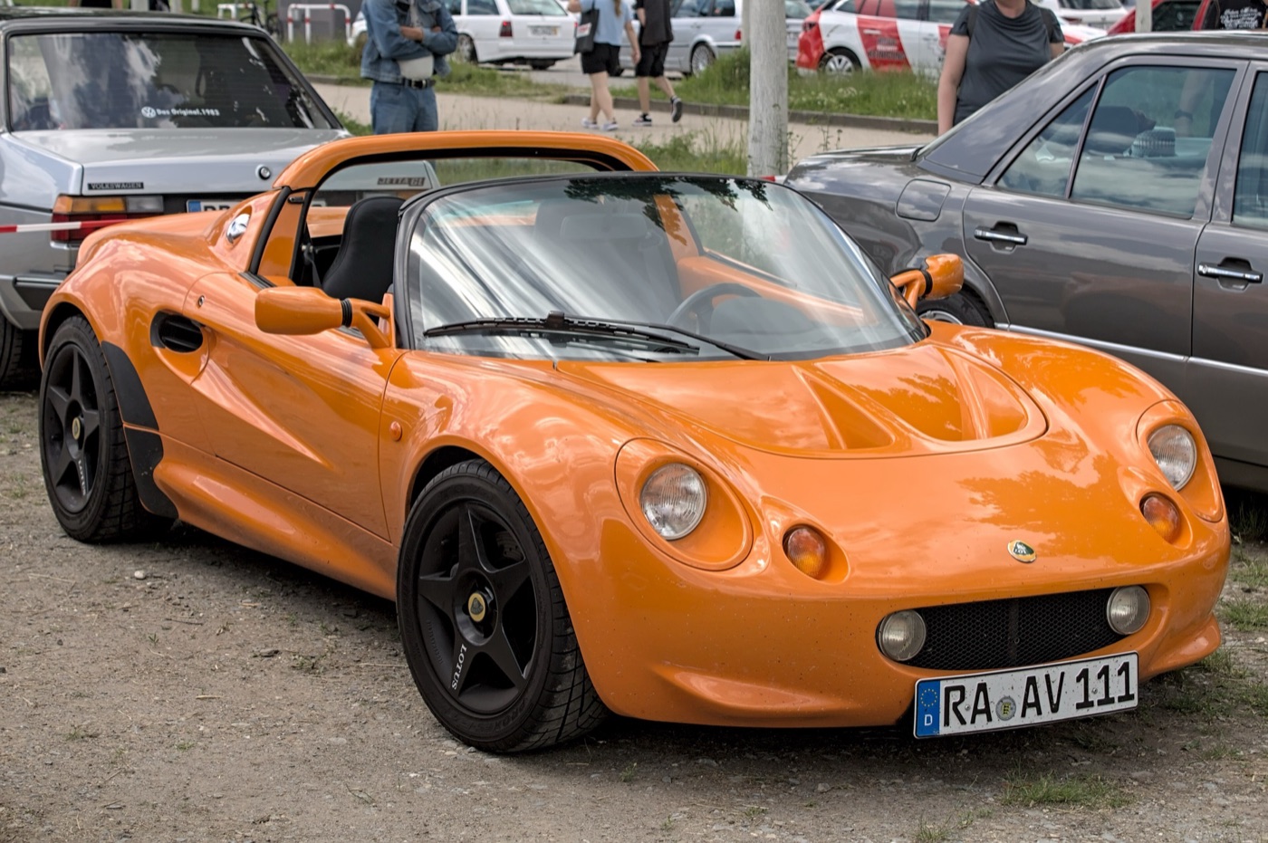 Orange first-series Lotus Elise roadster with black wheels parked among other cars