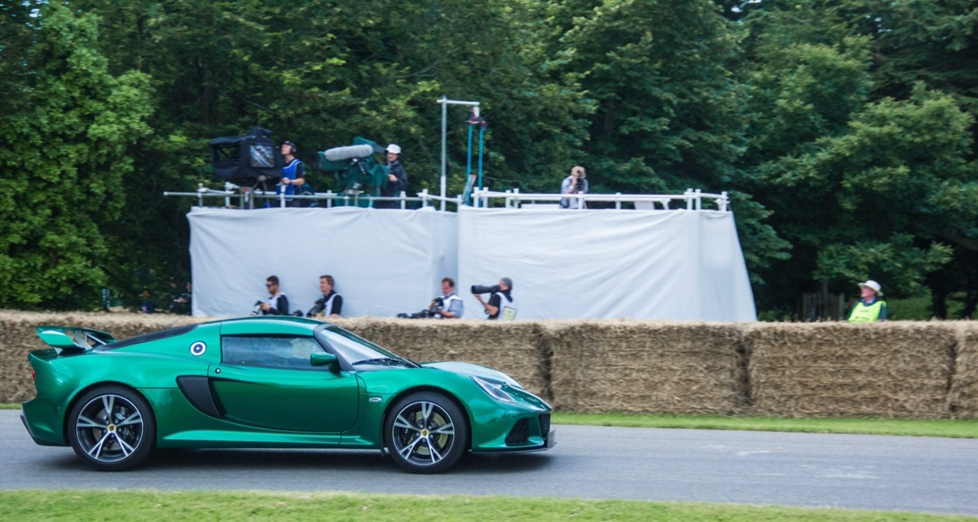 Green Lotus Exige S coupé driving past straw bales and photographers on a hill climb course