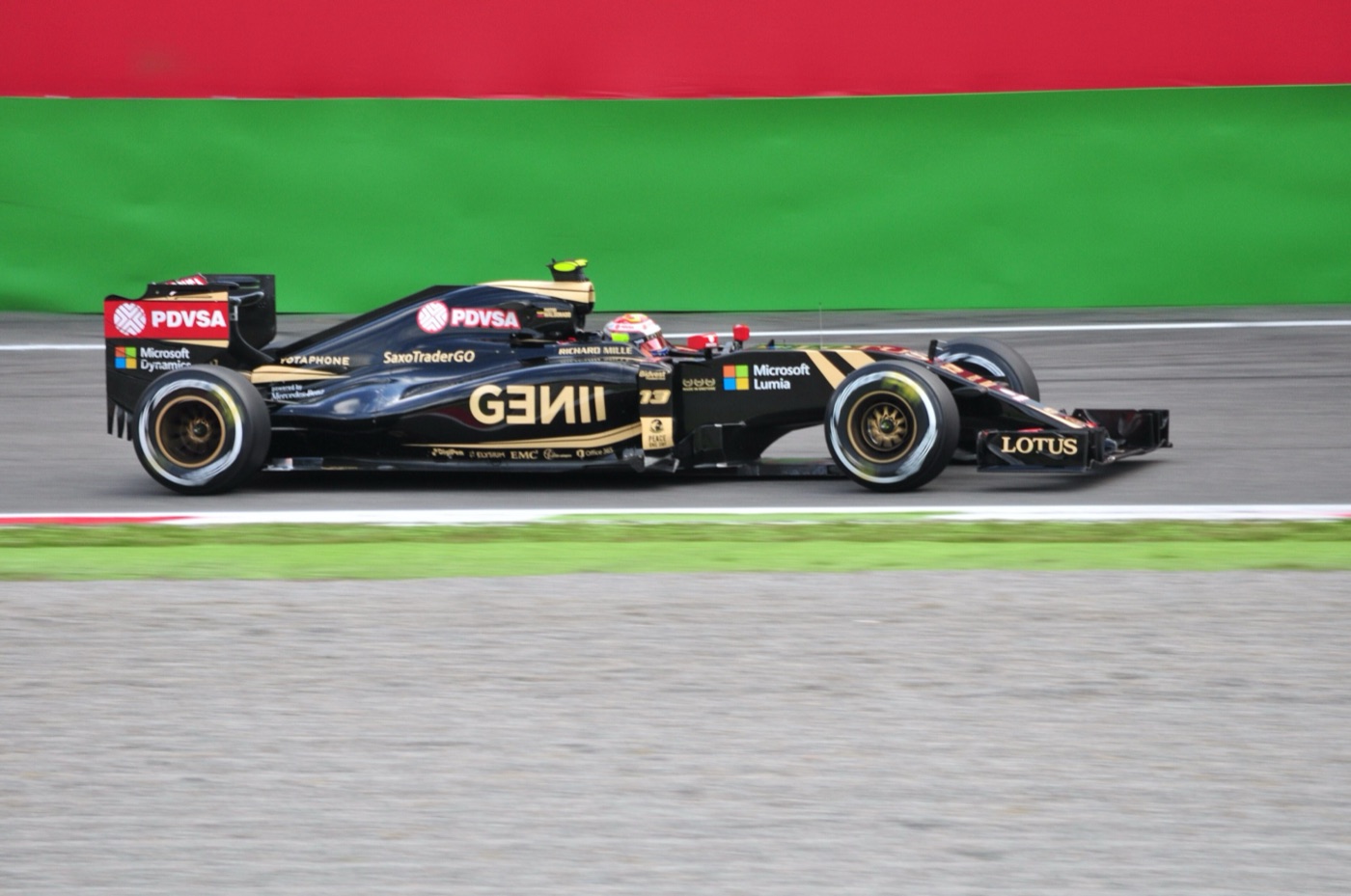 Black and gold Lotus E23 Hybrid with PDVSA and Microsoft logos at speed beside a red and green barrier