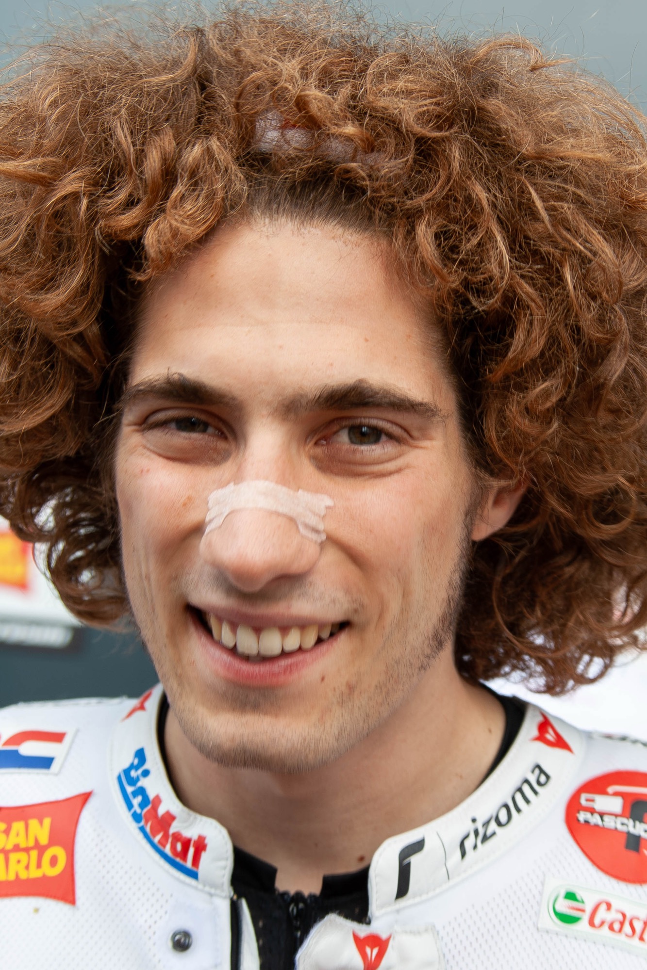 Head and shoulders portrait of Marco Simoncelli smiling, with his curly hair and a plaster across his nose, in San Carlo Gresini leathers