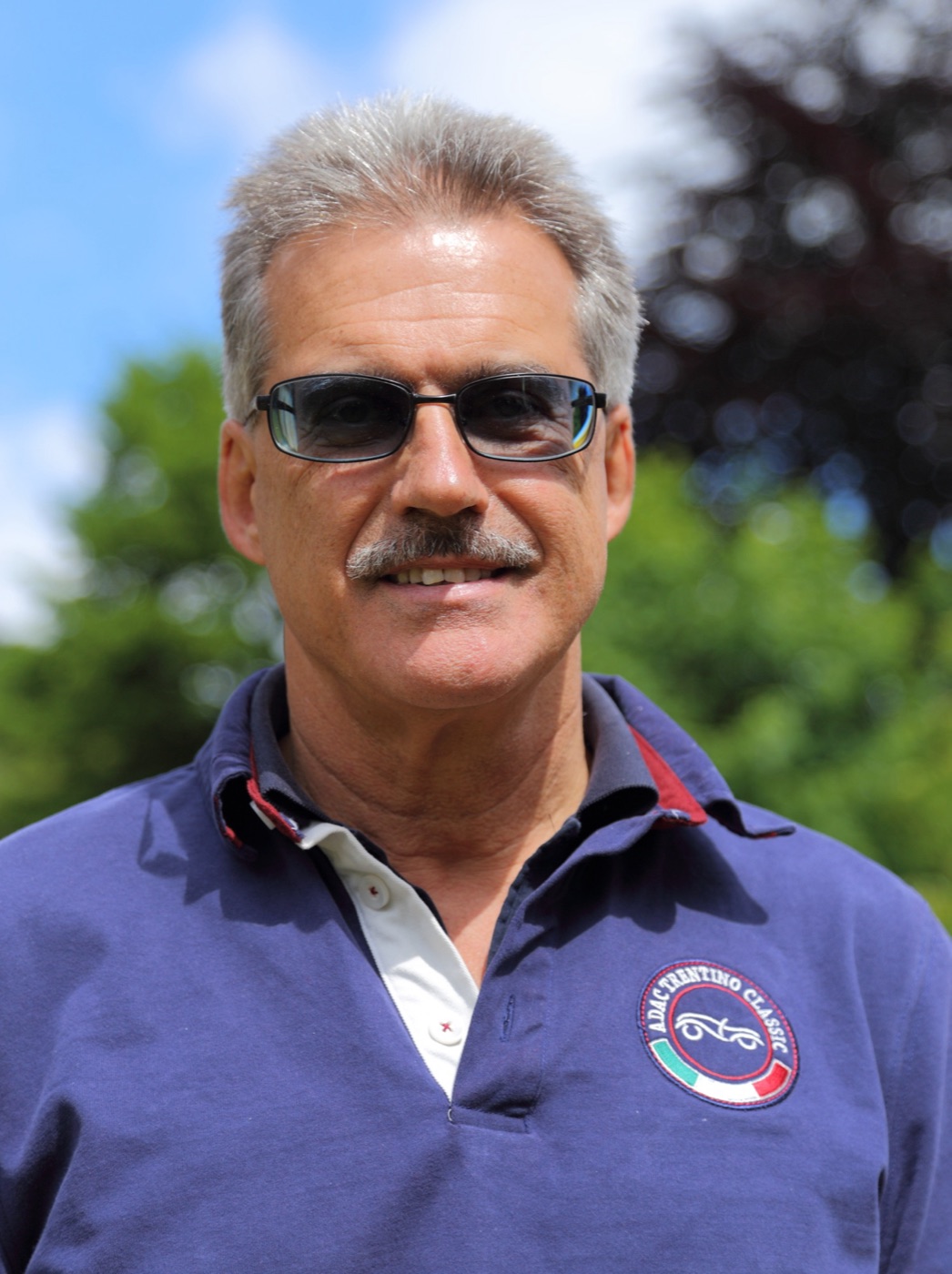 Mario Theissen, grey-haired with a moustache and tinted glasses, in a navy ADAC Trentino Classic polo shirt, outdoors in front of trees