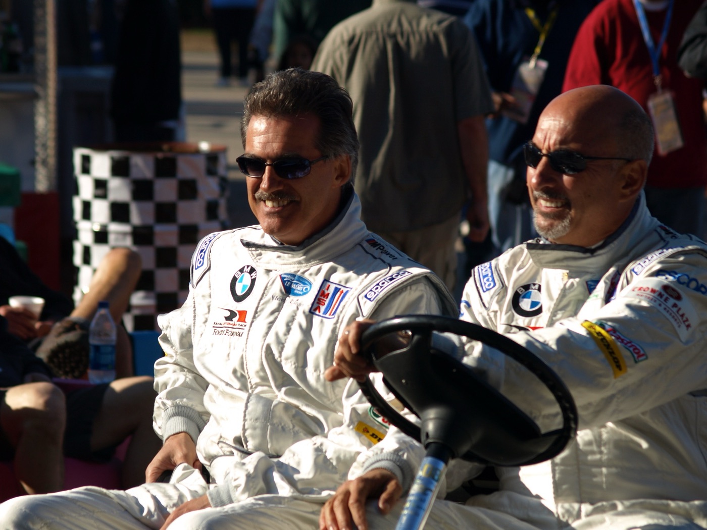 Mario Theissen and Bobby Rahal, both in white BMW race suits and sunglasses, riding a golf cart through a paddock