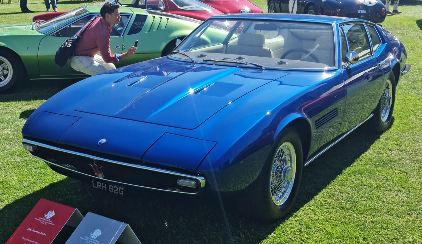 Blue 1969 Maserati Ghibli coupé with pop-up headlamps closed and wire wheels, parked on grass at a concours