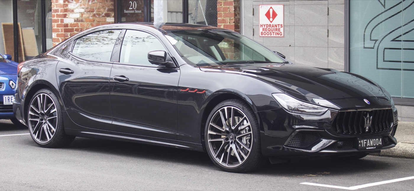 Black Maserati Ghibli Trofeo parked at a kerb, side-on front three-quarter view with large multi-spoke wheels and red-trimmed wing vents