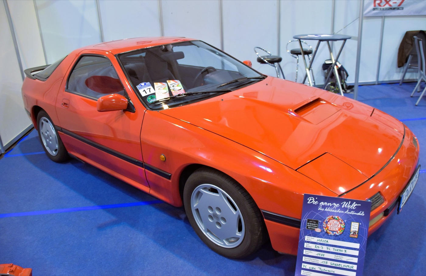Orange second-generation Mazda RX-7 Turbo II with a bonnet scoop, displayed indoors on a blue carpet
