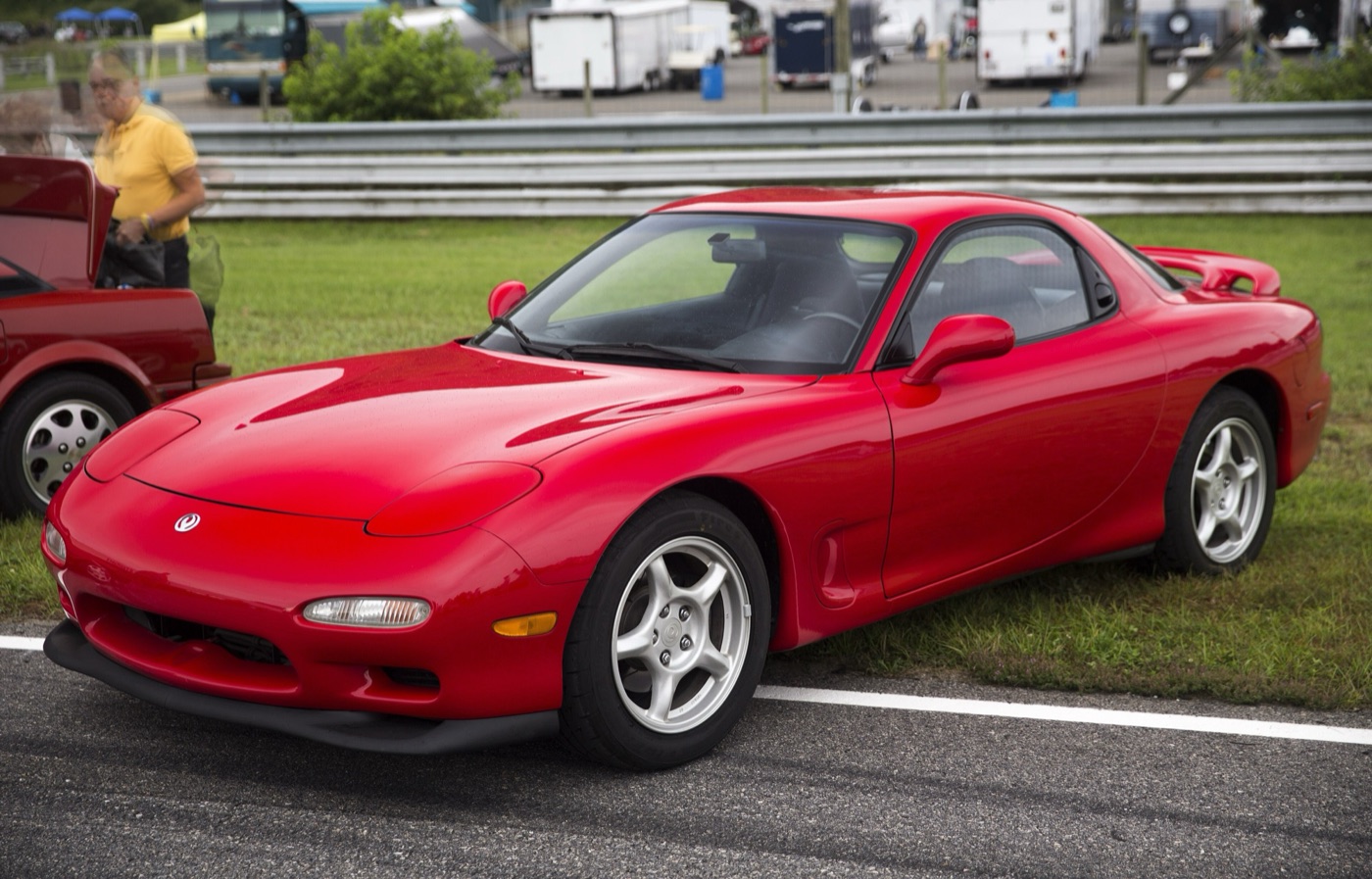 Red third-generation Mazda RX-7 with five-spoke wheels and a rear spoiler, parked on the grass beside a race track