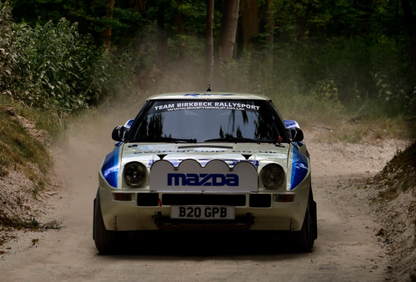 White and blue Mazda RX-7 Group B rally car driving through dust on a forest stage