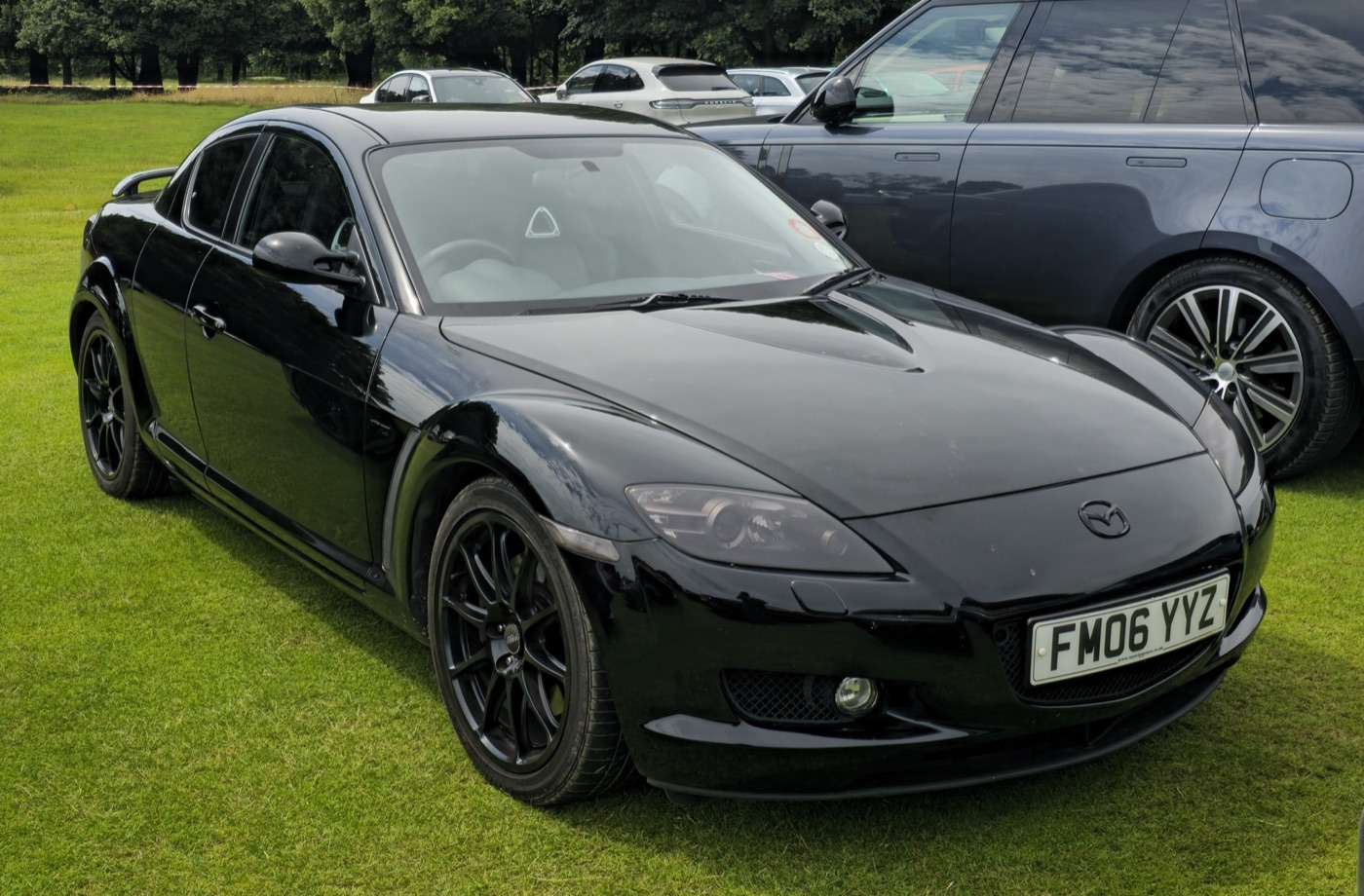 Black Mazda RX-8, identified by the photographer as a PZ, with black ten-spoke wheels and a rear spoiler, parked on grass among other cars
