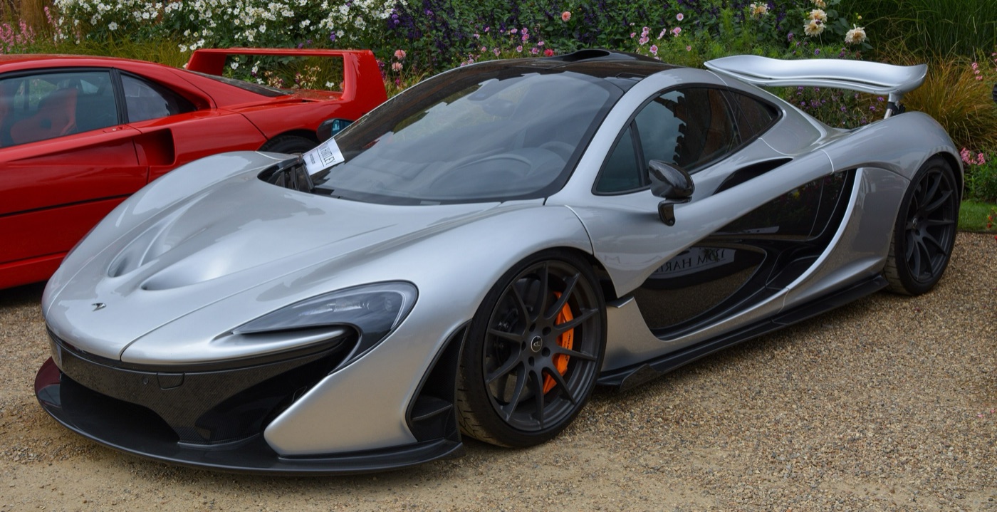 Silver McLaren P1 with its rear wing raised, parked on gravel at a concours beside a red Ferrari F40