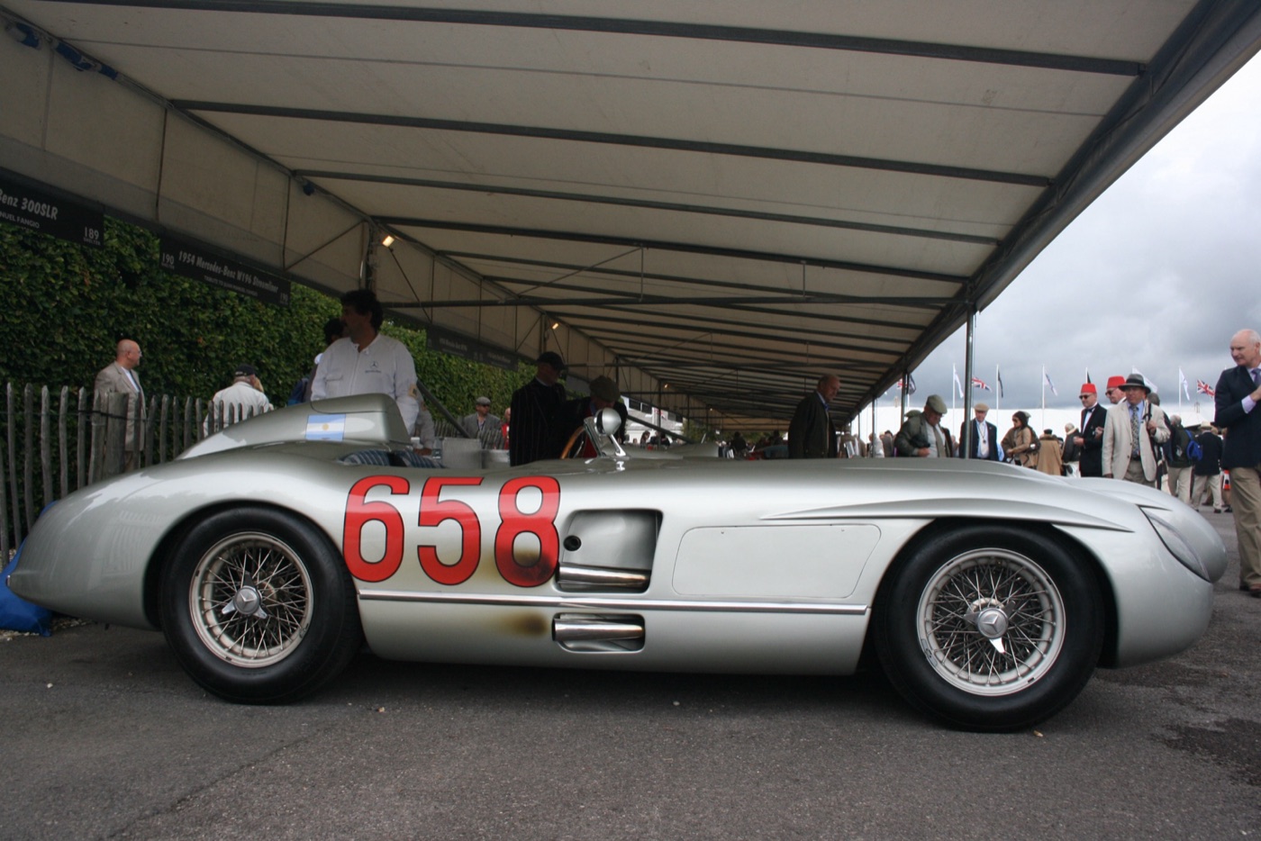 Side view of a silver Mercedes-Benz 300 SLR with the red race number 658 and side exhaust, parked under an awning