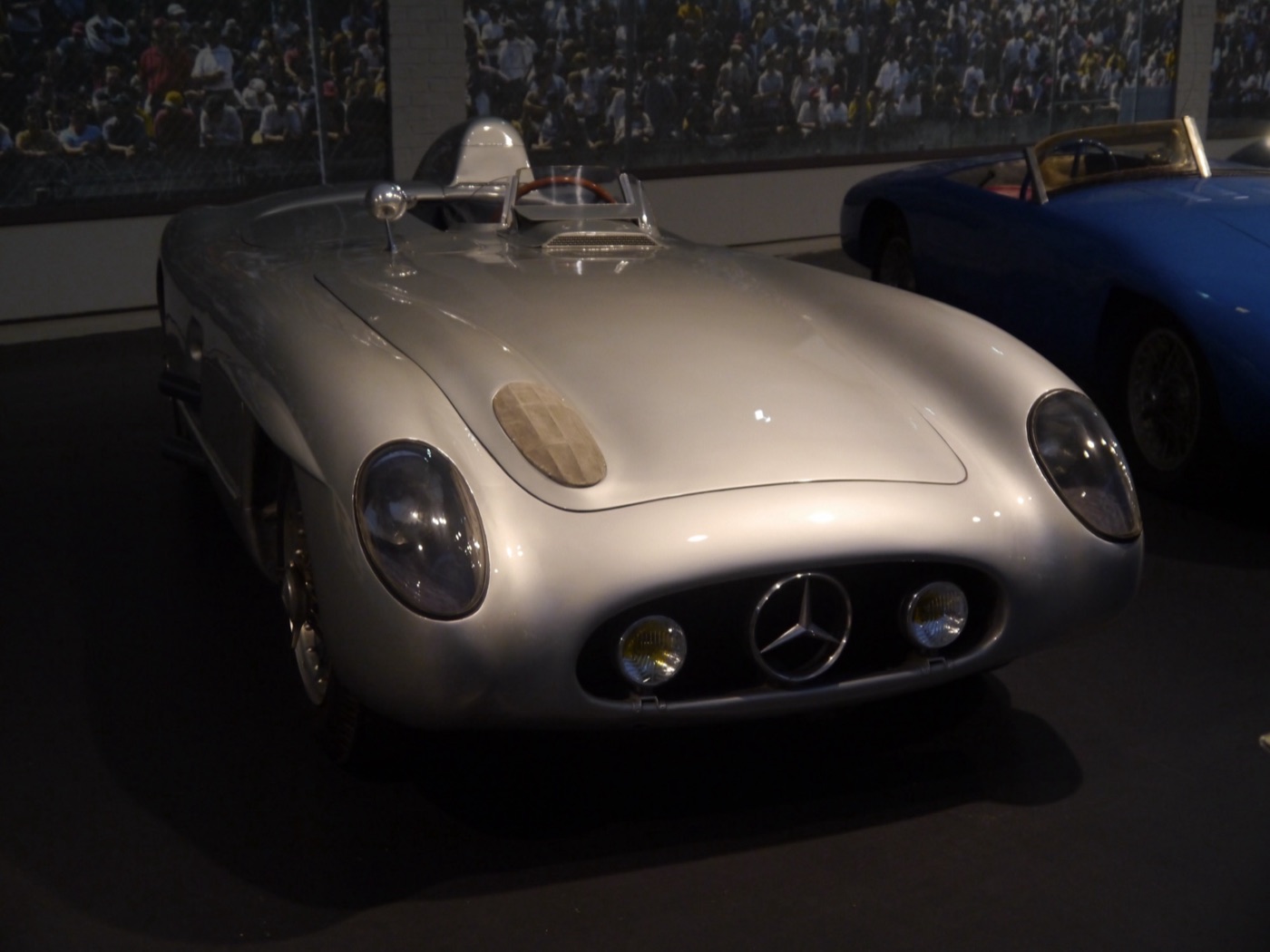 Silver Mercedes-Benz 300 SLR roadster with a headrest fairing, shown indoors in a museum next to a blue racing car