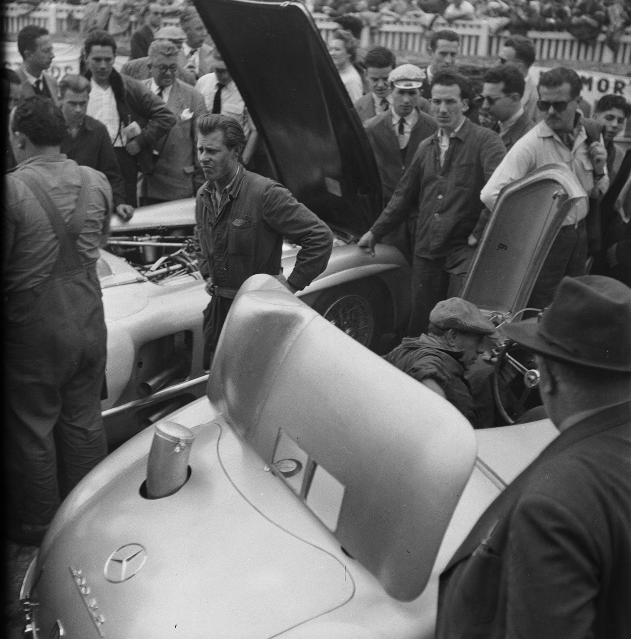 Black and white photo of mechanics and onlookers around Mercedes-Benz 300 SLRs in the pits at Le Mans, one with its large rear air-brake flap raised