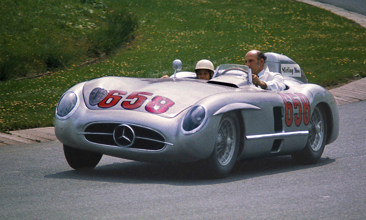 Stirling Moss driving the Mercedes-Benz 300 SLR numbered 658 with a passenger in a white helmet, on a track bordered by grass