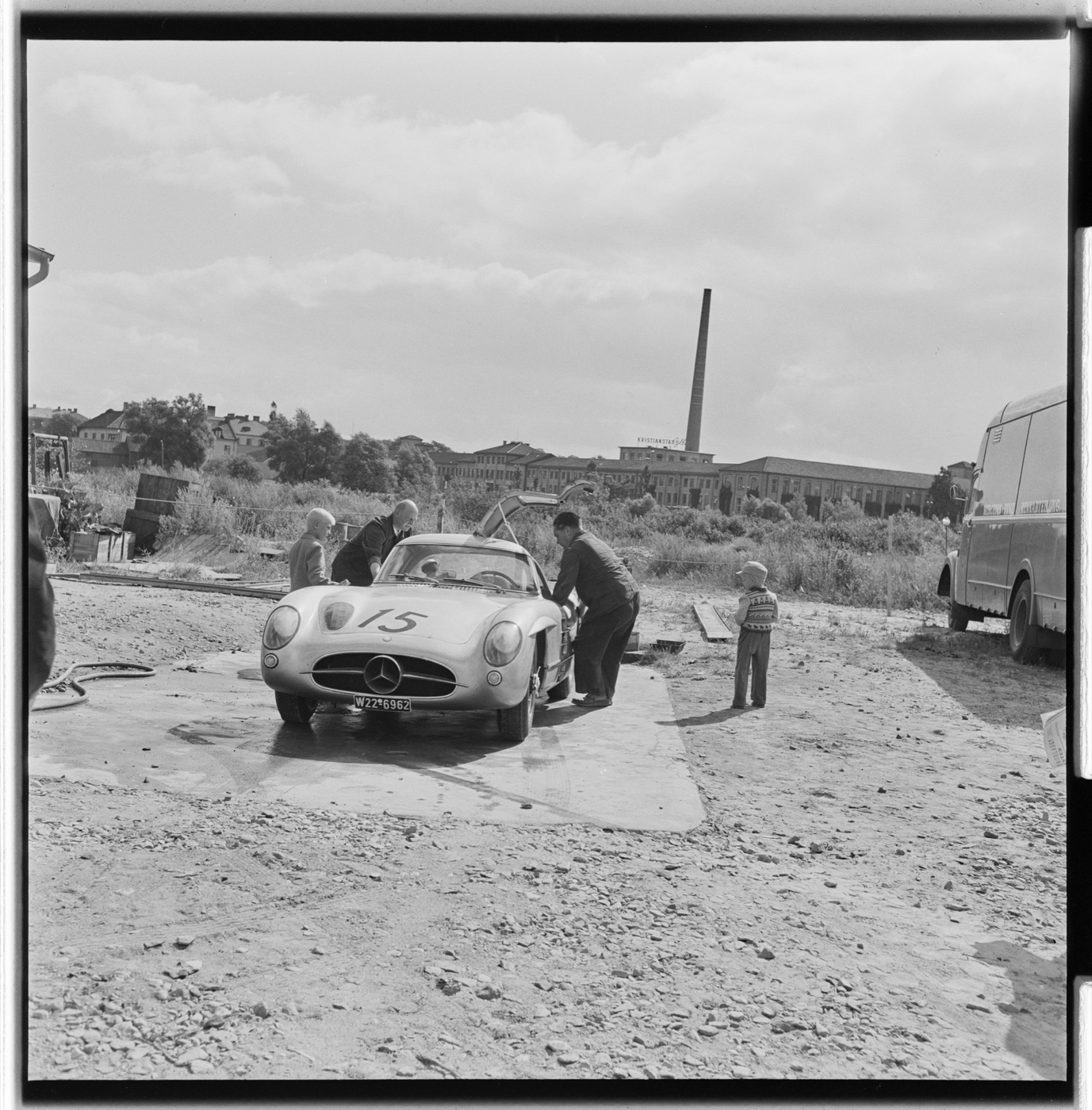 Black and white photo of a Mercedes-Benz 300 SLR coupé with race number 15 and German registration W22-6962, its gull-wing door open, on a gravel yard