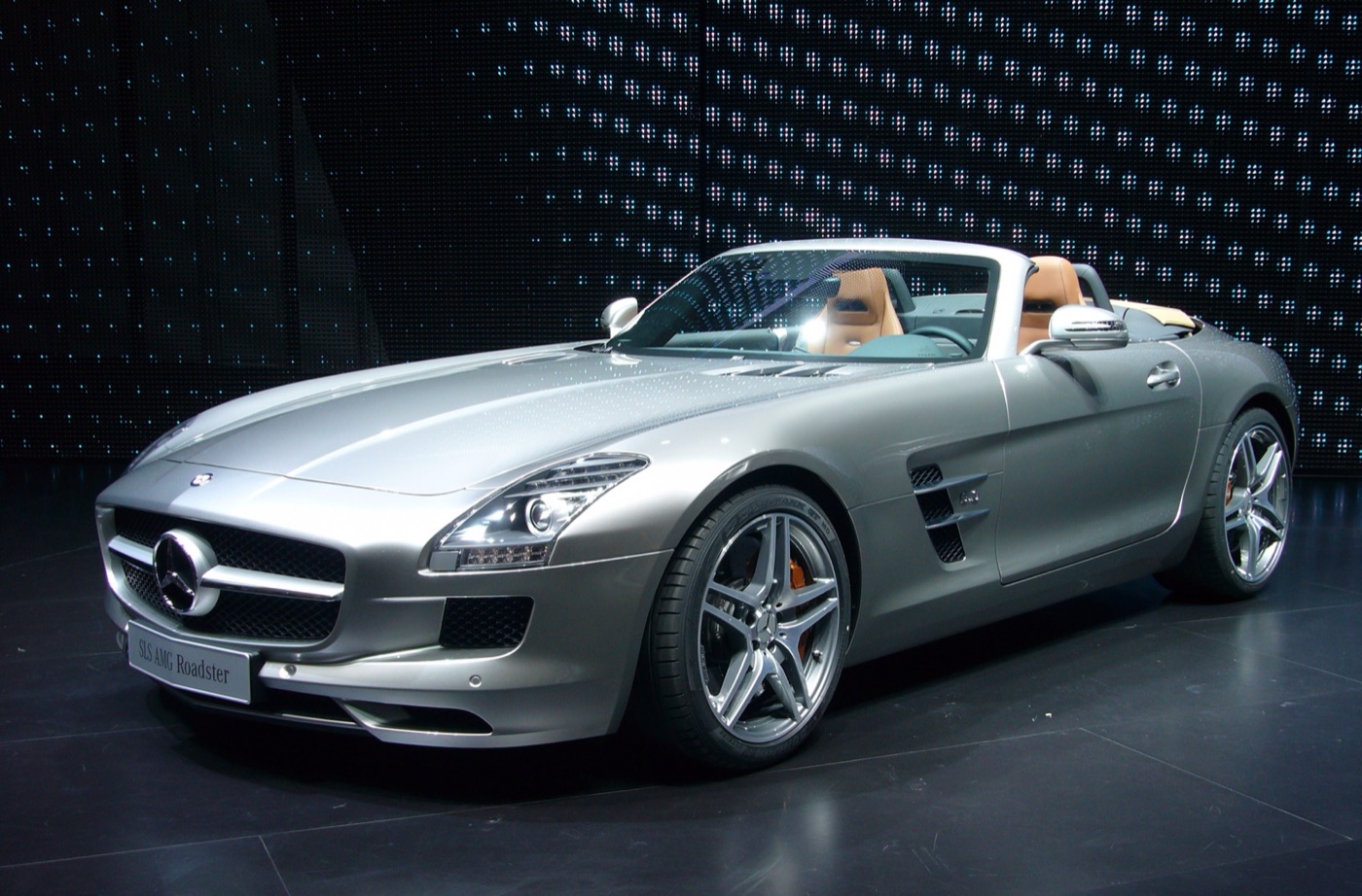Silver SLS AMG Roadster with its fabric roof folded and tan seats, on a dark motor show stand