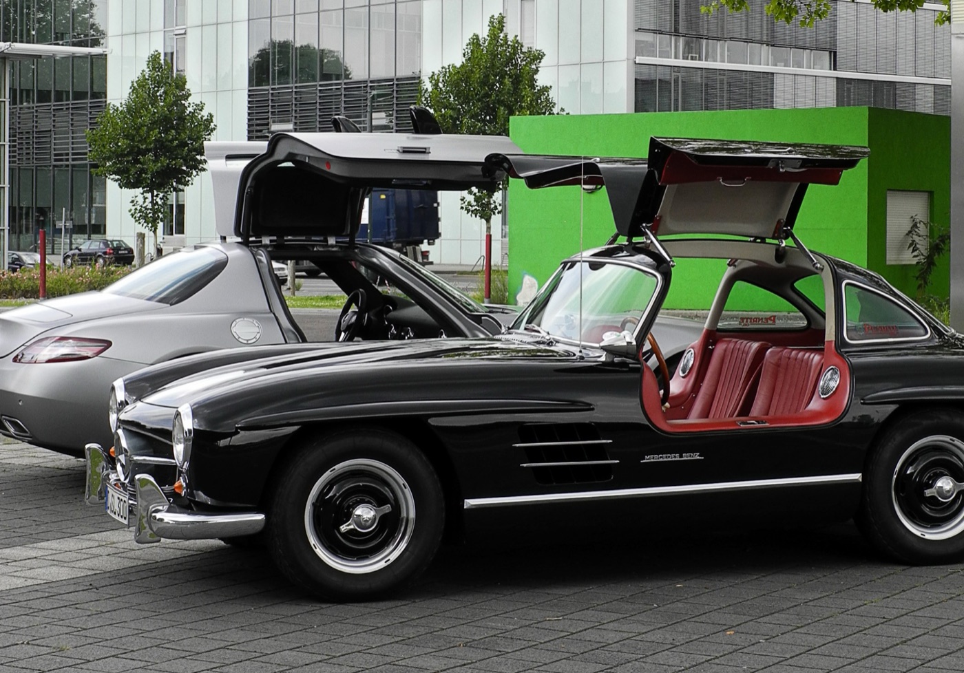 Black 1950s Mercedes 300 SL Gullwing with red interior and a silver SLS AMG behind it, the doors of both cars raised
