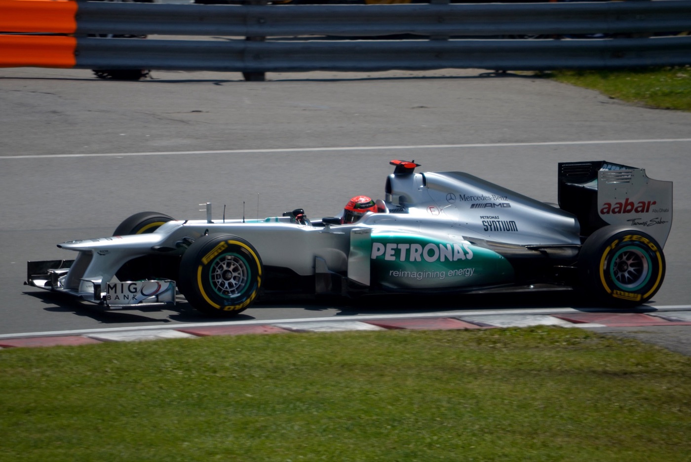 Silver and turquoise Mercedes Formula One car with yellow-marked Pirelli tyres, driver in a red helmet, beside a red and white kerb and grass