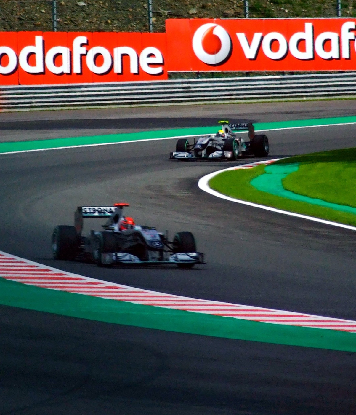 Two silver Mercedes Formula One cars on a curving track in front of red Vodafone advertising boards, the leading driver in a red helmet