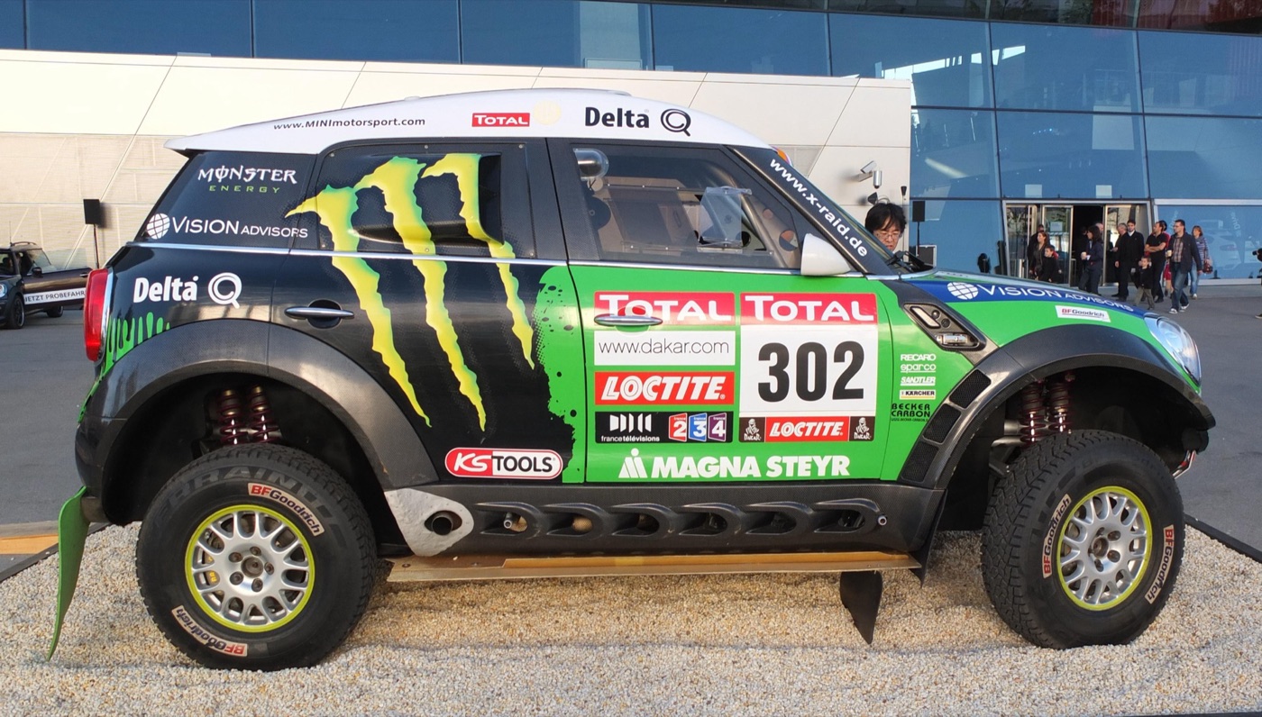 Green, black and white MINI ALL4 Racing number 302 in Monster Energy livery, displayed on gravel outside a glass building