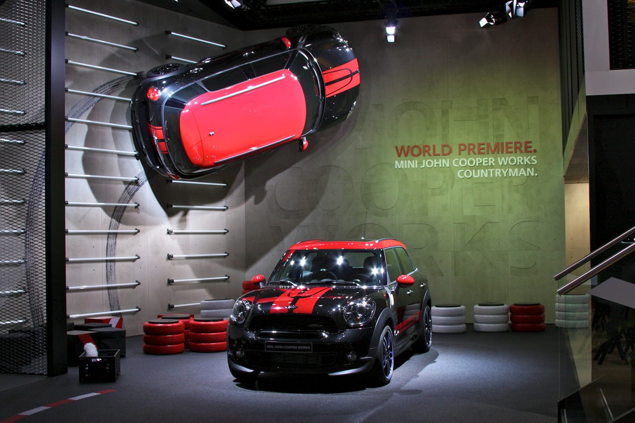 Black MINI John Cooper Works Countryman with red roof and bonnet stripes on a stand under a World Premiere sign, with a second car mounted on the wall above