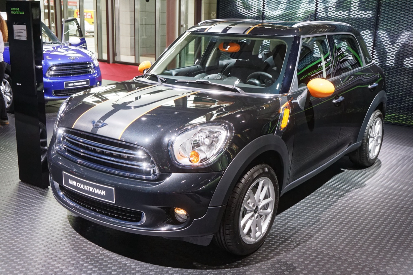 Dark grey facelifted MINI Countryman with grey bonnet stripes and orange mirror caps on a show stand