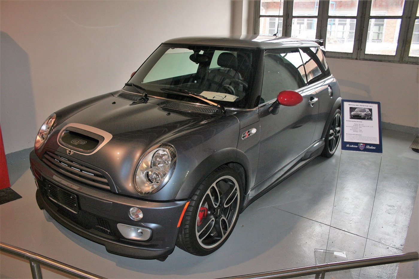 Grey 2006 MINI Cooper S John Cooper Works GP with red mirror caps on display inside a museum hall beside an information board