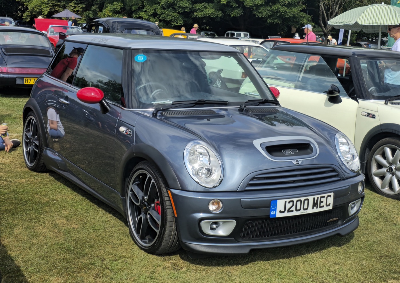 Grey-blue 2006 MINI Cooper S with John Cooper Works GP Kit with red mirror caps and black four-spoke wheels parked on grass at a car show