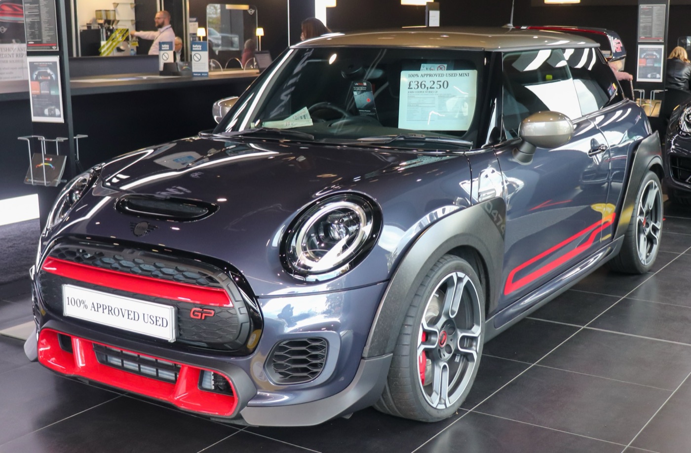 Racing Grey MINI John Cooper Works GP with red front grille trim in a dealer showroom, with a price card reading £36,250 in the windscreen