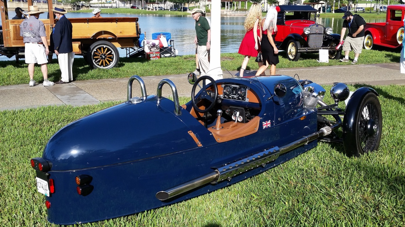 Dark blue 2012 Morgan 3 Wheeler seen from the rear quarter on grass, showing its tapering tail, twin roll hoops, tan leather cockpit and side exhaust